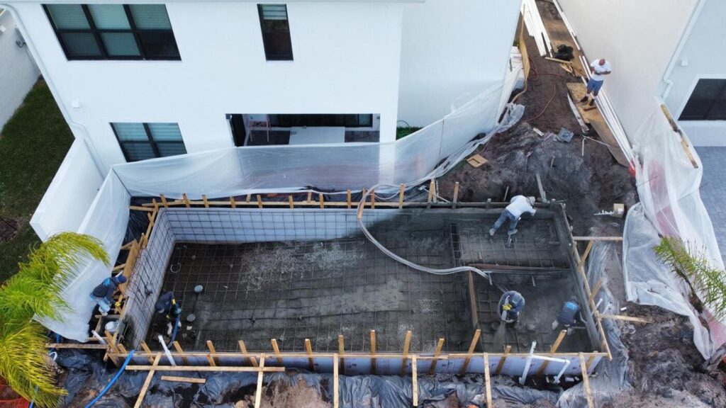 Common Mistakes to Avoid When Hiring a Pool Builder 2 Aerial view of a pool construction site between two houses, with workers applying shotcrete to the rebar frame.