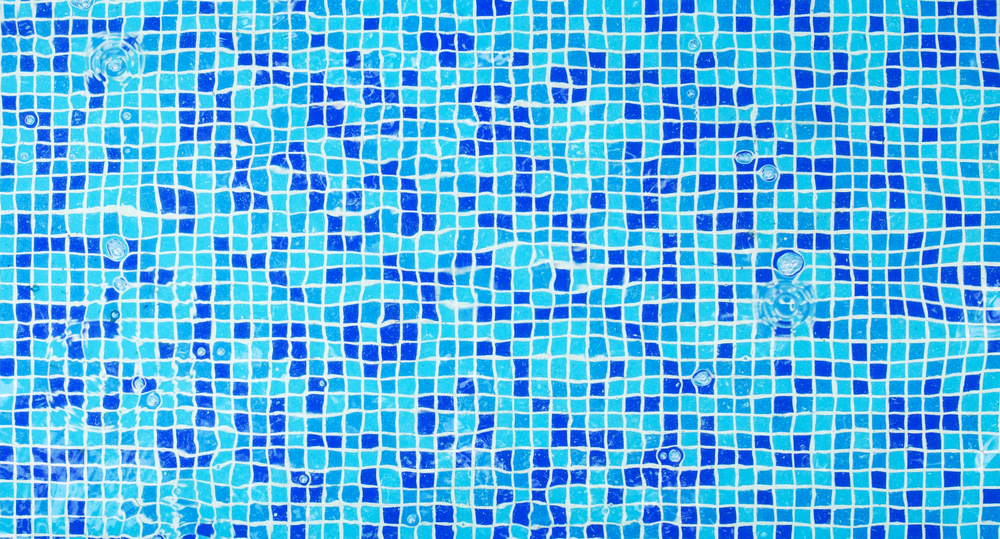 A close-up of a swimming pool's blue mosaic tile bottom, visible through the rippling water.