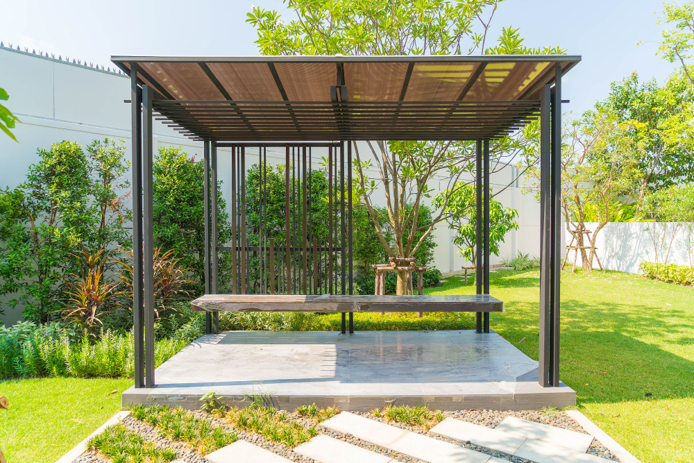 A modern black metal pergola with a built-in wooden bench on a stone patio surrounded by a lush green lawn and garden.