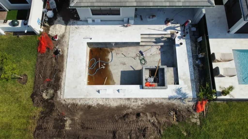 Common Mistakes to Avoid When Hiring a Pool Builder 1 An aerial shot of a backyard under construction, showing a newly poured concrete pool shell and patio, with dirt and construction equipment visible.