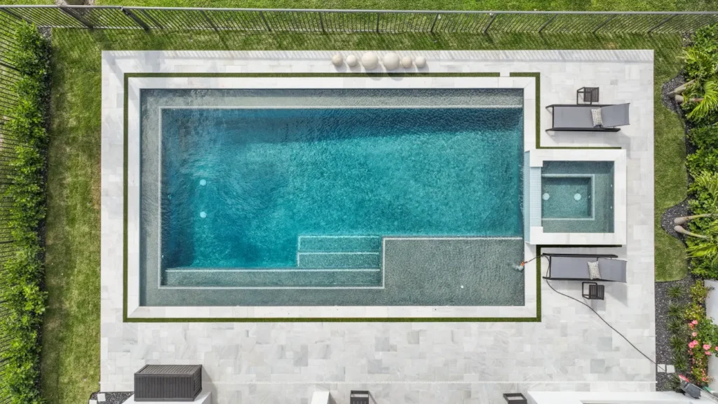 Finding the Perfect Fit: Choosing the Right Pool Builder for You 3 An aerial view of a newly remodeled backyard pool and raised spa, featuring a sun shelf with in-pool loungers and a gray tile patio.