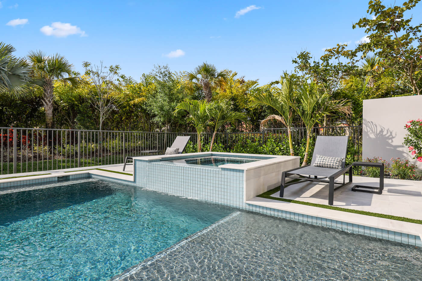 A modern swimming pool with a raised jacuzzi, sun shelf, and decorative tile accents, with two lounge chairs on the patio and tropical landscaping in the background.