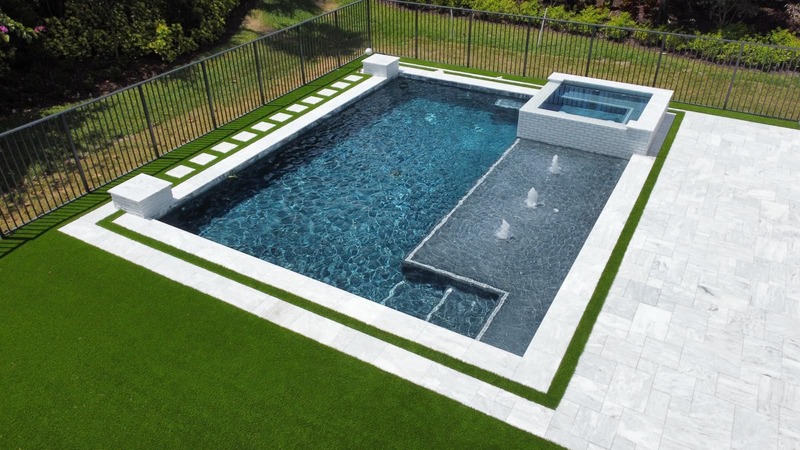 A modern in-ground pool with a raised jacuzzi, decorative fountains, and a stone patio surrounded by artificial turf.