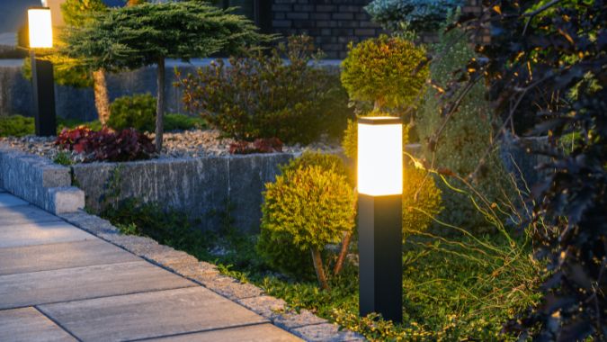 A modern, square-shaped outdoor lamp post illuminating a landscaped garden path with pavers and various plants.