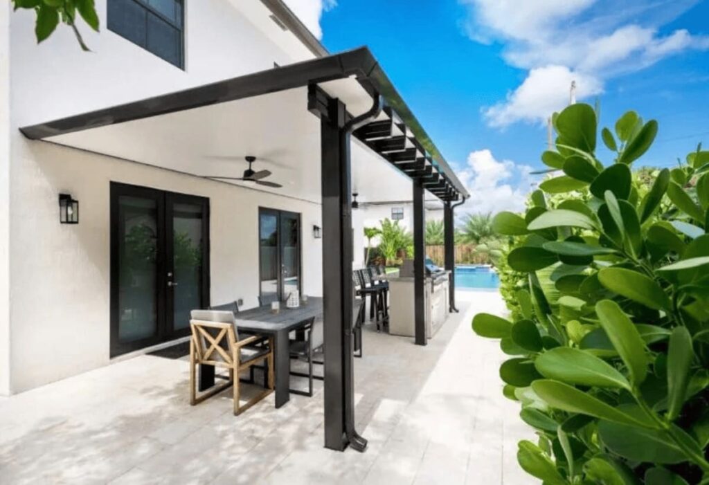 Finding Your Perfect Fit: Choosing the Right Pergola Installer 3 A modern outdoor living space featuring a white house with a black-and-white patio cover. Underneath, there's a dining table with chairs and a built-in outdoor kitchen with a grill. A pool is visible in the background.