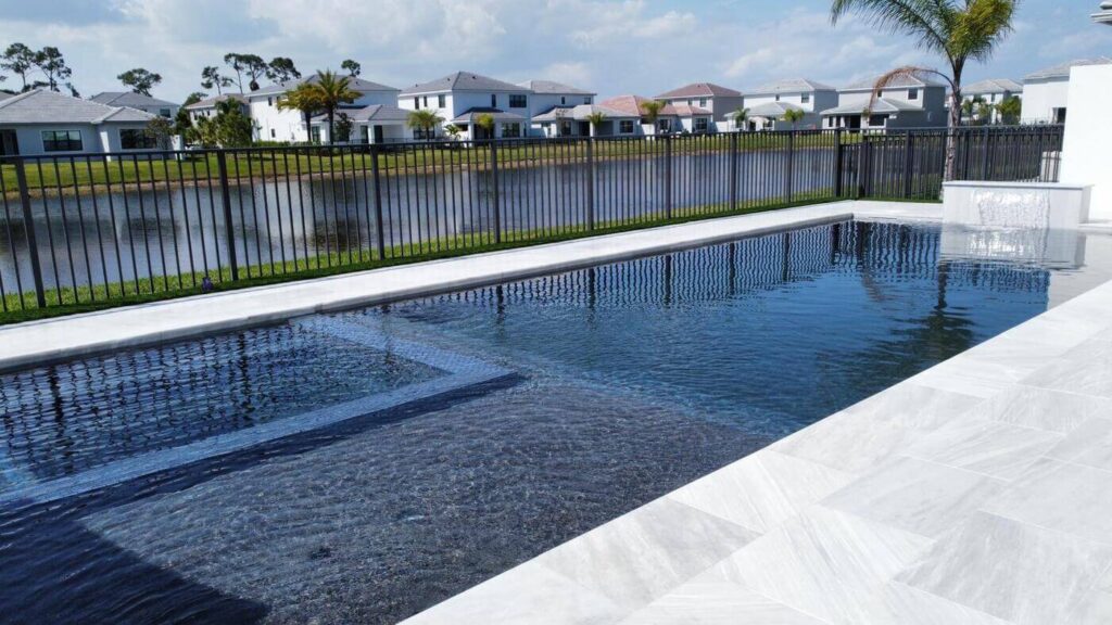 Finding the Perfect Fit: Choosing the Right Pool Builder for You 1 A side view of a modern rectangular pool and patio area that overlooks a lake bordered by a black metal fence.