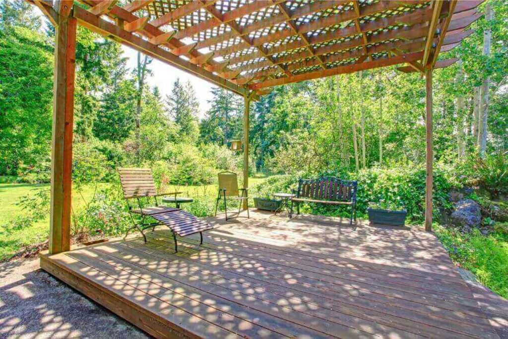 Finding Your Perfect Fit: Choosing the Right Pergola Installer 2 rustic wooden pergola with a latticed roof, covering a small wooden deck. The deck holds various outdoor chairs and a bench, surrounded by a lush, green, natural landscape.