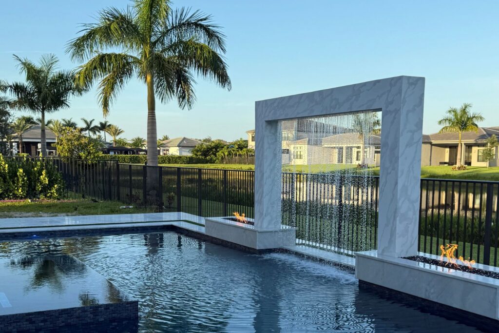 Luxury backyard pool with modern custom water features, including a waterfall wall and fire elements, surrounded by tropical landscaping for a relaxing outdoor space.