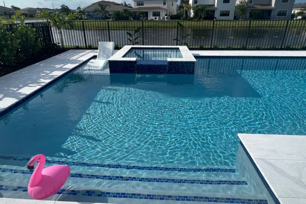 Luxury Florida pool with a Baja shelf and in-water chaise lounge chairs, surrounded by modern tile decking, clear blue water, and lush tropical landscaping for a stylish backyard retreat.