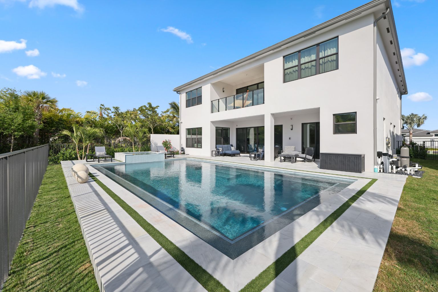 Modern luxury Florida backyard with a rectangular swimming pool and integrated spa, surrounded by a clean white tile deck, lounge seating, and tropical landscaping, showcasing a contemporary pool design with glass railing balcony view and clear blue sky
