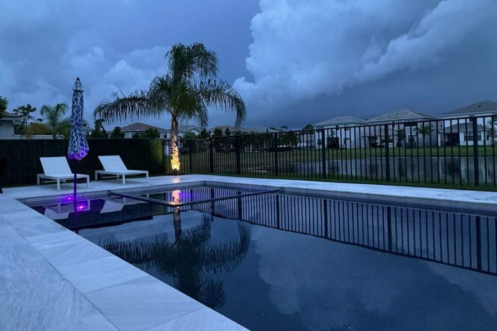 Florida backyard with a modern swimming pool, white pool deck, lounge chairs, and umbrella, captured just before an approaching storm with dark clouds in the sky reflecting on the water surface.