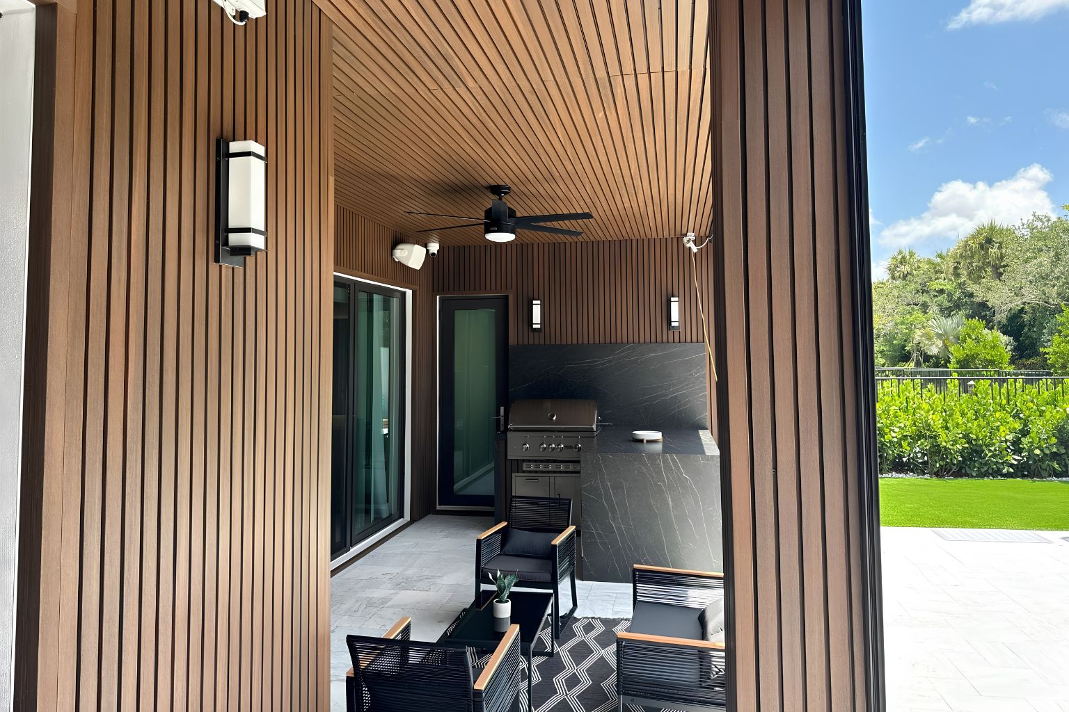 Modern outdoor kitchen with wood-paneled patio, built-in grill, sleek black furniture, and seamless flow to backyard pool, ideal for South Florida luxury home entertaining