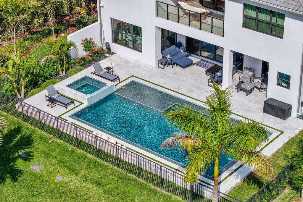 Aerial view of a modern backyard pool in Florida, featuring a rectangular pool with integrated spa, lounge chairs, patio seating, surrounded by lush landscaping and a safety fence, showcasing a completed luxury pool construction.