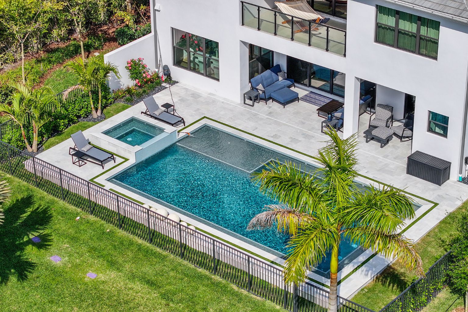 Aerial view of a modern backyard pool in Florida, featuring a rectangular pool with integrated spa, lounge chairs, patio seating, surrounded by lush landscaping and a safety fence, showcasing a completed luxury pool construction.