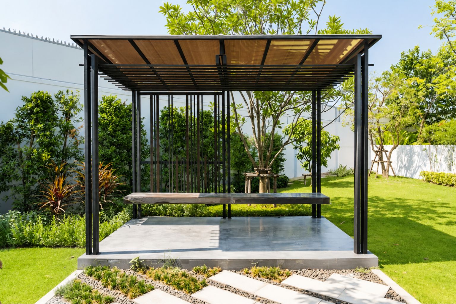 Freestanding hurricane-rated pergola with black frame, shaded bench, concrete patio, and landscaped backyard garden.