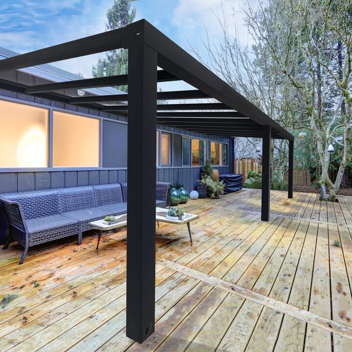 A black aluminum pergola structure installed on a rustic wooden deck, providing shade over an outdoor seating area with a gray woven sofa.
